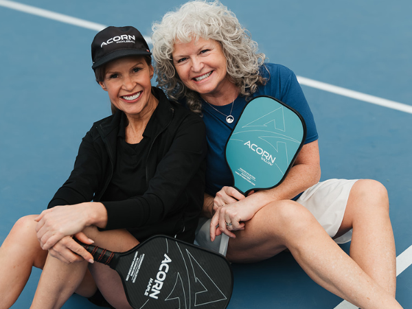 Two women smiling, sitting on a court with Acorn Pickleball paddles